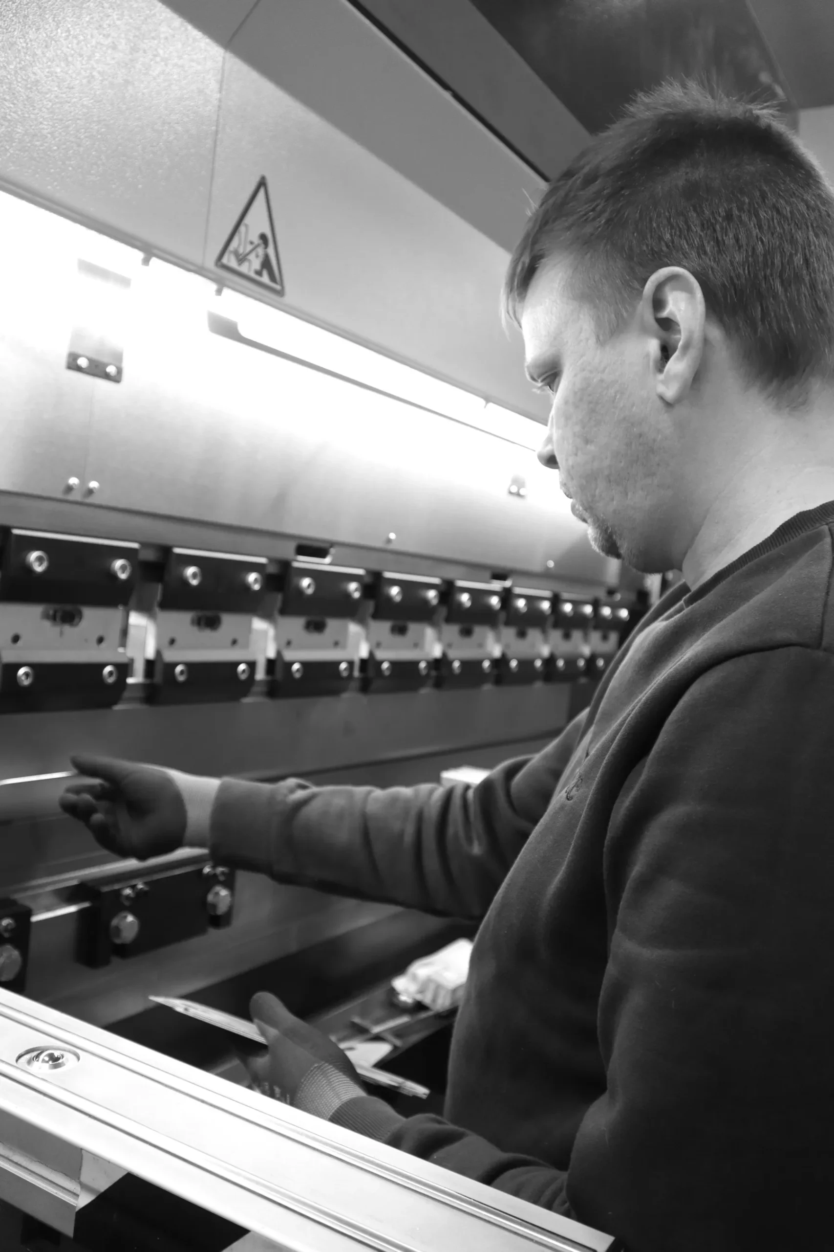 En man i sweatshirt manövrerar en stor industrimaskin tillverkad av borstat rostfritt stål och justerar spakar med vänster hand. Den svartvita bilden fångar atmosfären i verkstaden och anspelar på klassisk svensk design.