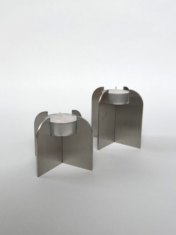 Två små vita värmeljus är placerade i moderna, minimalistiska silverfärgade metallhållare med korsande böjda paneler. Ljusstakarna Ljusstakar - Värmeljus har en geometrisk design och är placerade på en slät vit bakgrund.