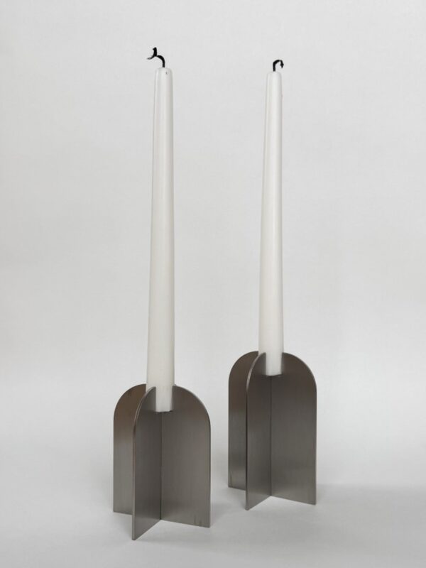 Två Ljusstakar - Antikljus vita stearinljus med brända vekar sitter i moderna, geometriska metallhållare med korsande platta paneler, placerade mot en vit bakgrund.